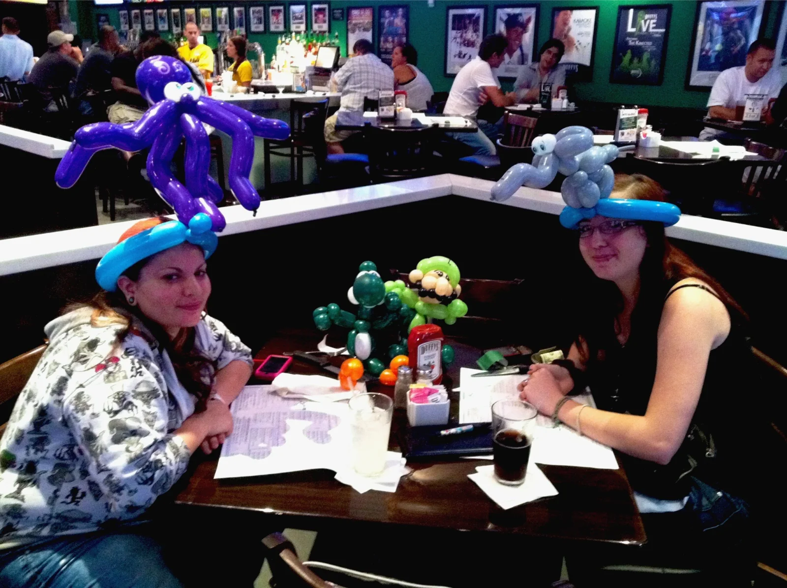 Two adult restaurant guests wearing elaborate balloon hats at their table.