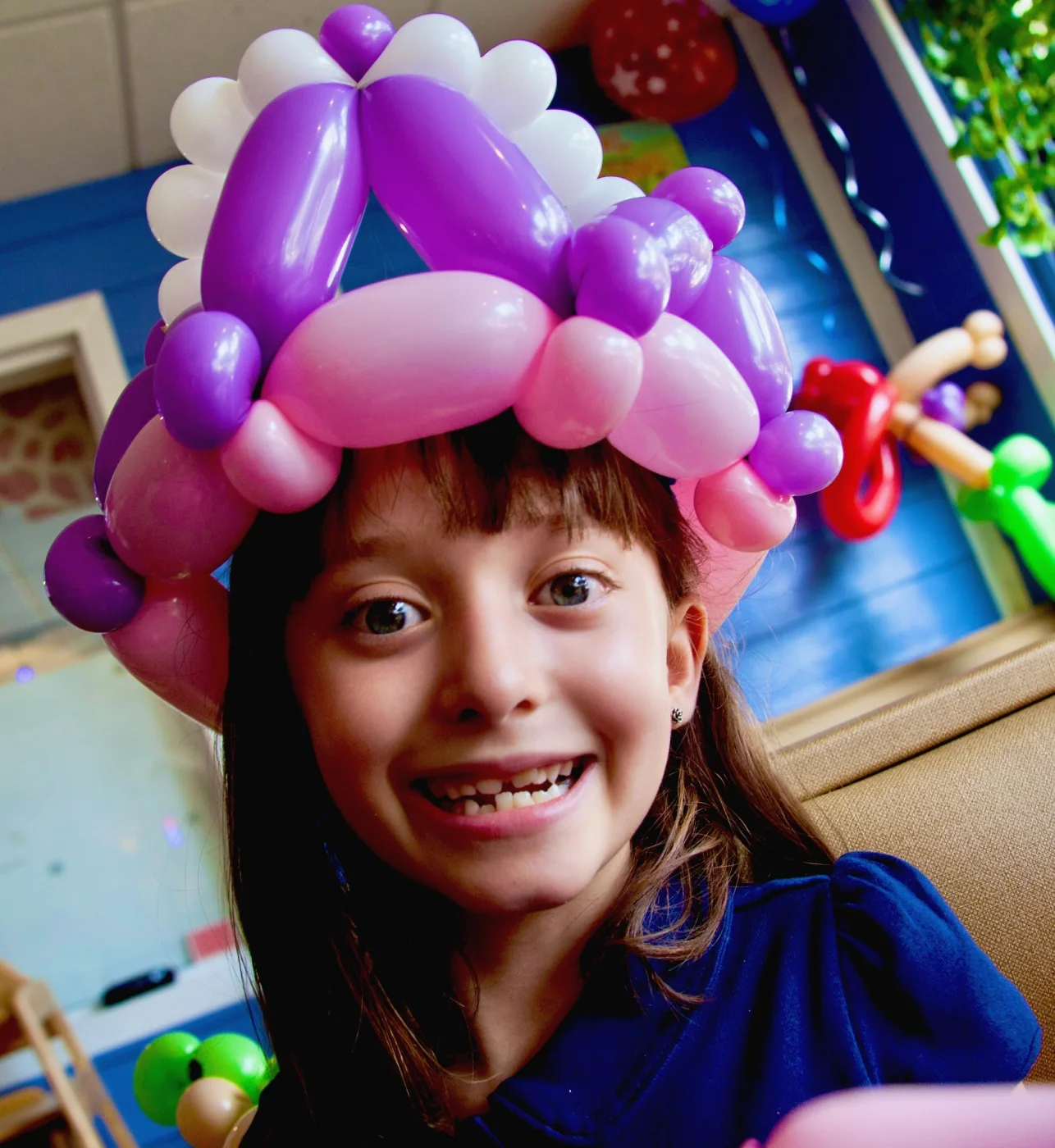 A smiling girl wearing an elaborate purple and pink balloon crown.