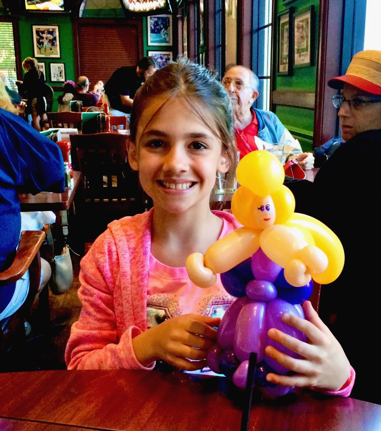 A girl at a restaurant table holding a purple balloon sculpture.