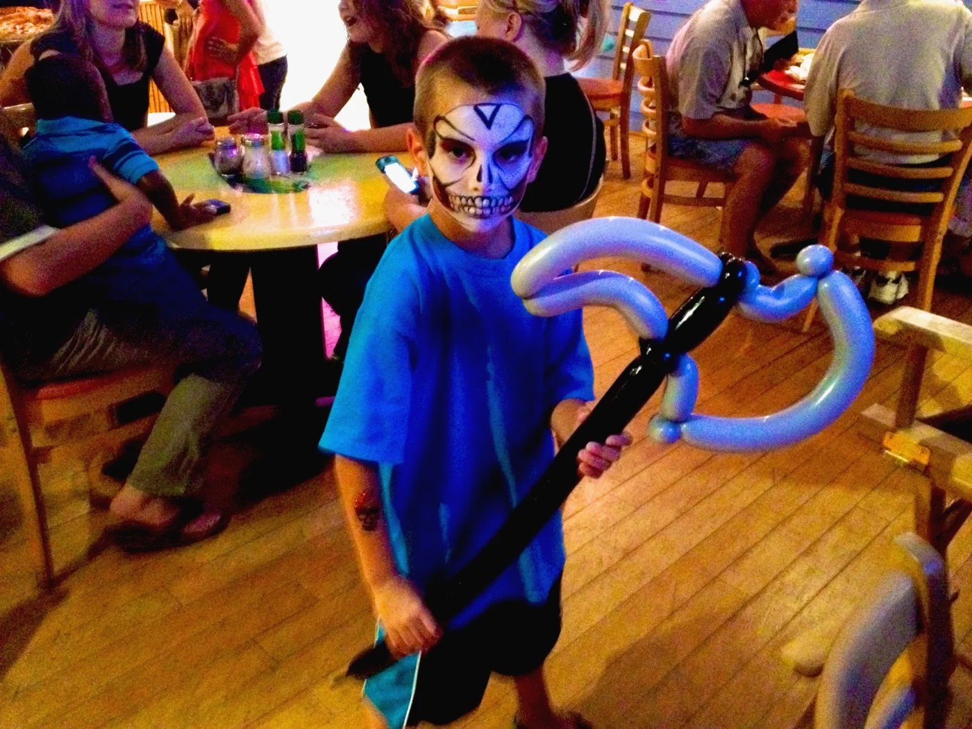 A boy in a restaurant holding a large blue balloon creation while wearing skull face paint.