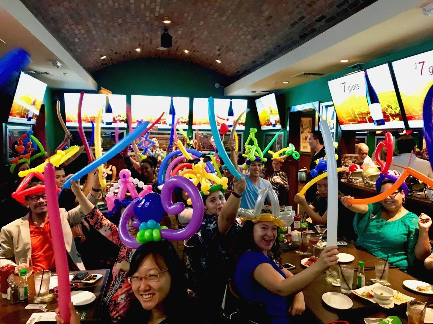 Restaurant guests raising colorful balloon hats and balloon swords around their tables.
